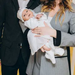 Elegancko ubrani rodzice trzymają na rękach niemowlę w białym ubranku. Idealny przykład, jak ubrać się na chrzest wnuczki.