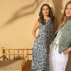 Dwie kobiety pozują obok bambusowego krzesła. Jedna w sukience w liście, druga w zielonej kurtce. Dowiedz się, jak zmierzyć sukienkę na płasko.