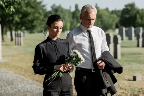 Smutna kobieta w czarnym stroju na pogrzeb trzyma kwiaty, obok mężczyzna w garniturze.