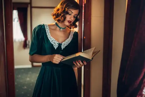 Kobieta w sukience w stylu lat 40. z koronkowym kołnierzykiem czyta książkę w zabytkowym wnętrzu.