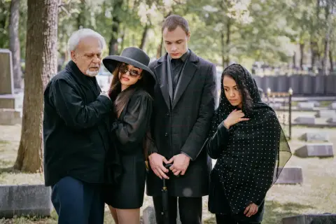 Smutna rodzina na cmentarzu. Elegancki ubiór damski na pogrzebie zimą, z kapeluszem i okularami przeciwsłonecznymi, obok żałobników w czerni.