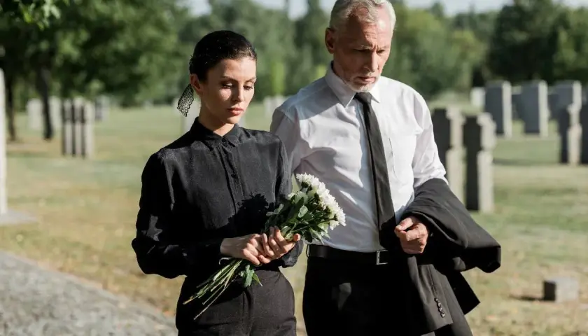 Smutna kobieta w czarnym stroju na pogrzeb trzyma kwiaty, obok mężczyzna w garniturze.