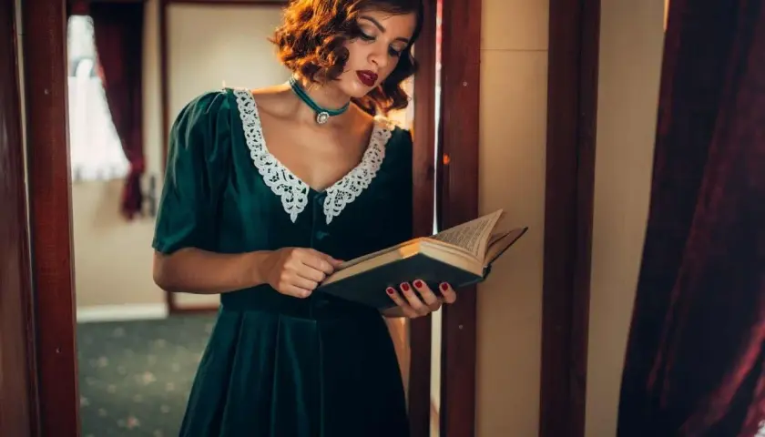 Kobieta w sukience w stylu lat 40. z koronkowym kołnierzykiem czyta książkę w zabytkowym wnętrzu.