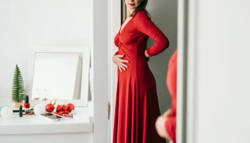 Kobieta w czerwonej, eleganckiej sukience, która optycznie wyszczupla. Na toaletce świąteczne ozdoby i kosmetyki.