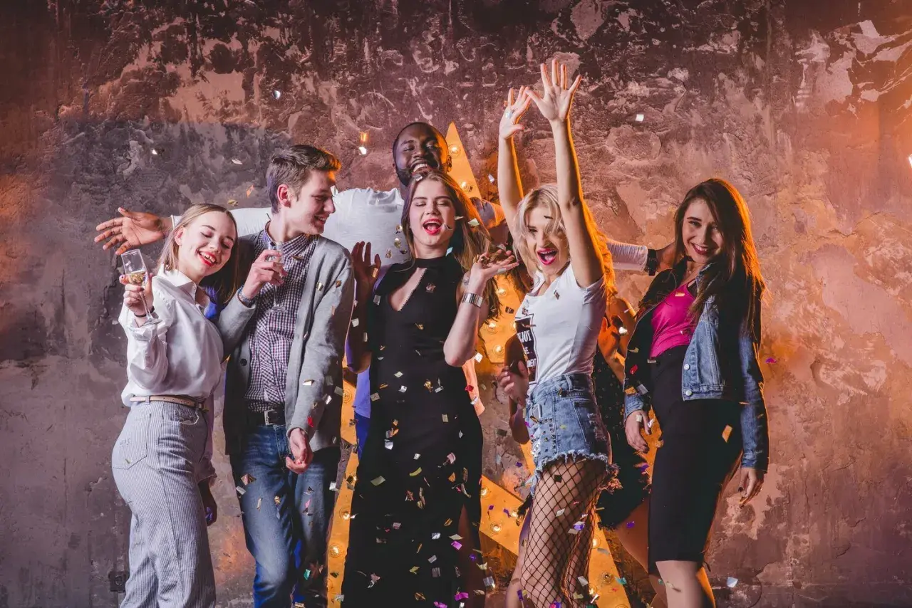 Grupa przyjaciół świętuje w klubie, sypiąc konfetti. Inspiracja, jak się ubrać do klubu: sukienka, jeansy, topy, marynarka.