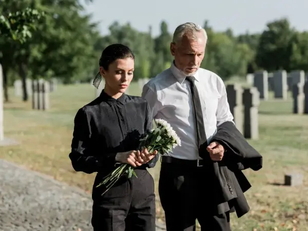 Smutna kobieta w czarnym stroju na pogrzeb trzyma kwiaty, obok mężczyzna w garniturze.